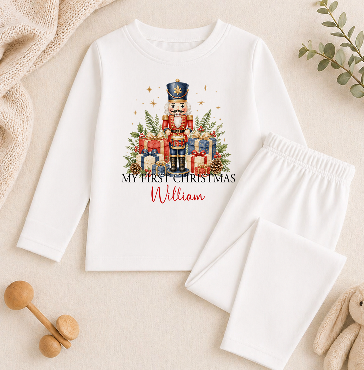A white long-sleeved shirt with a Christmas-themed design, including a nutcracker figure and presents, along with a pair of white pajama pants. The shirt also has the text "My first Christmas William" printed on it.
