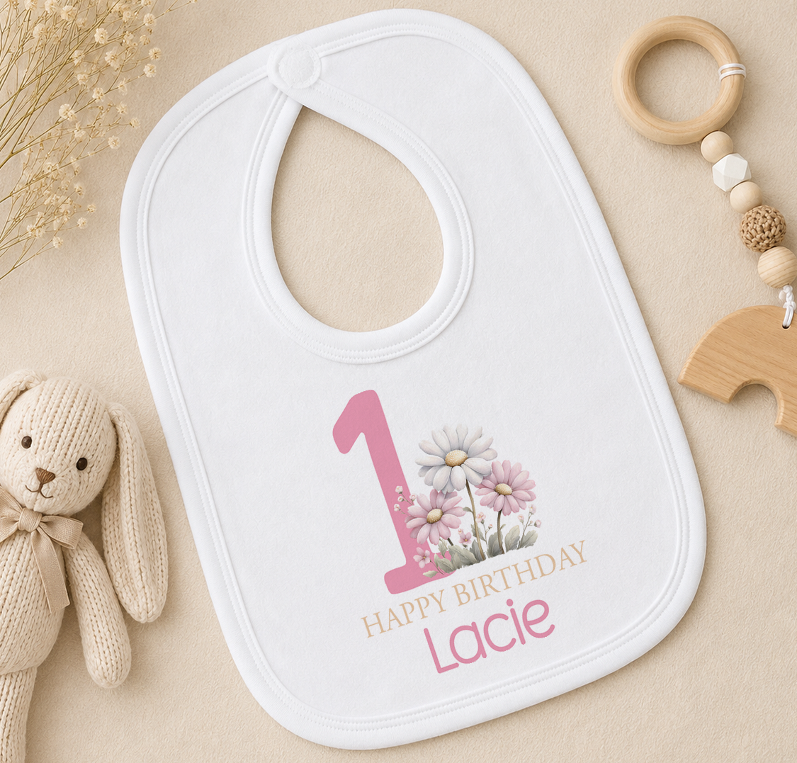 Personalised white bib with pink number one and floral 1st birthday design, styled with a wooden teether ring
