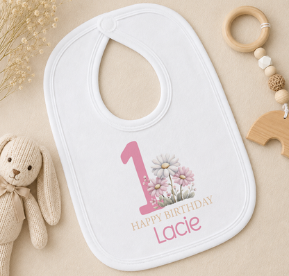 Personalised white bib with pink number one and floral 1st birthday design, styled with a wooden teether ring
