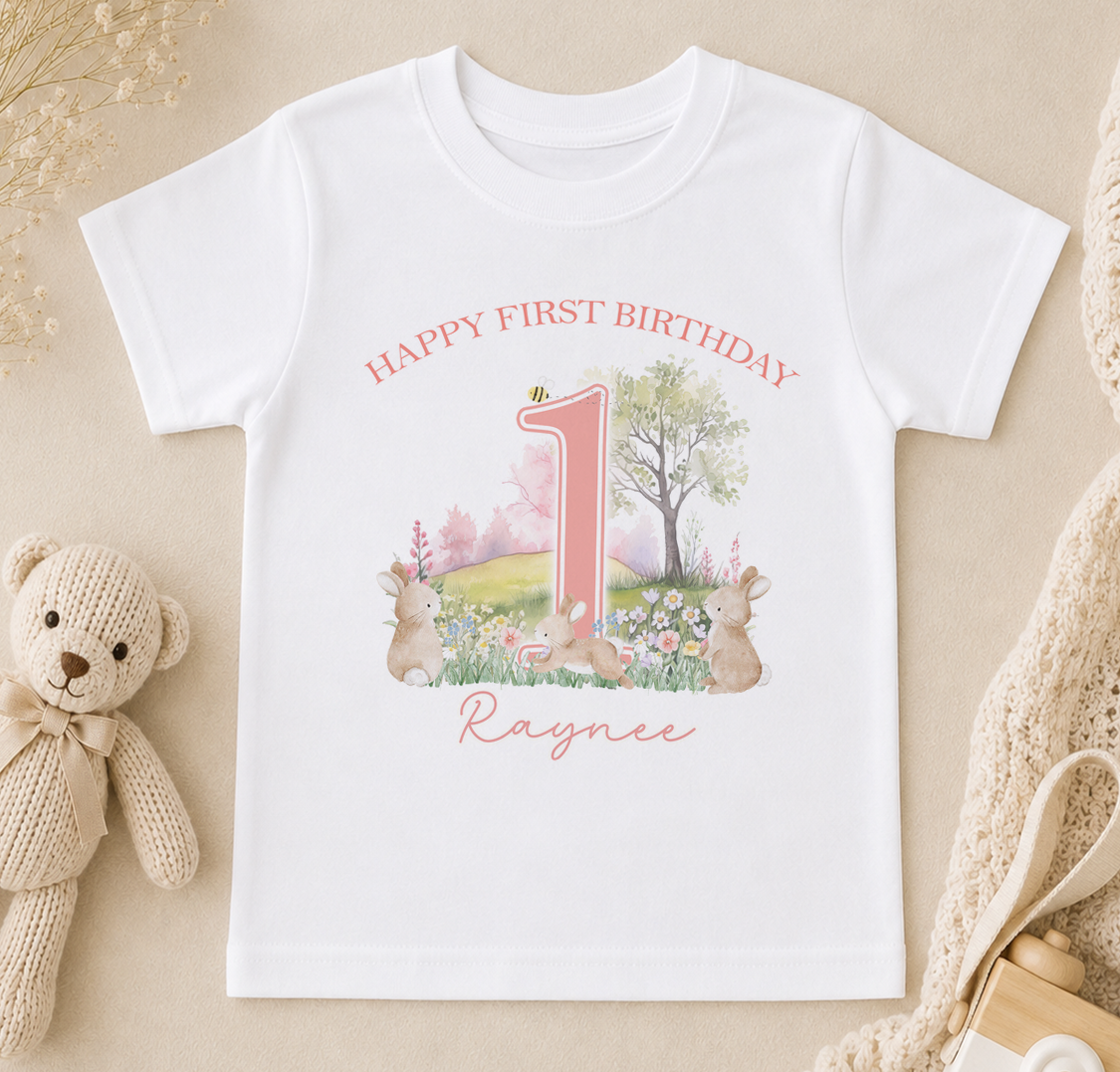 A white t-shirt with a pink number one and a garden scene printed on it, along with the text "Happy first birthday Raynee".