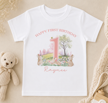 A white t-shirt with a pink number one and a garden scene printed on it, along with the text "Happy first birthday Raynee".