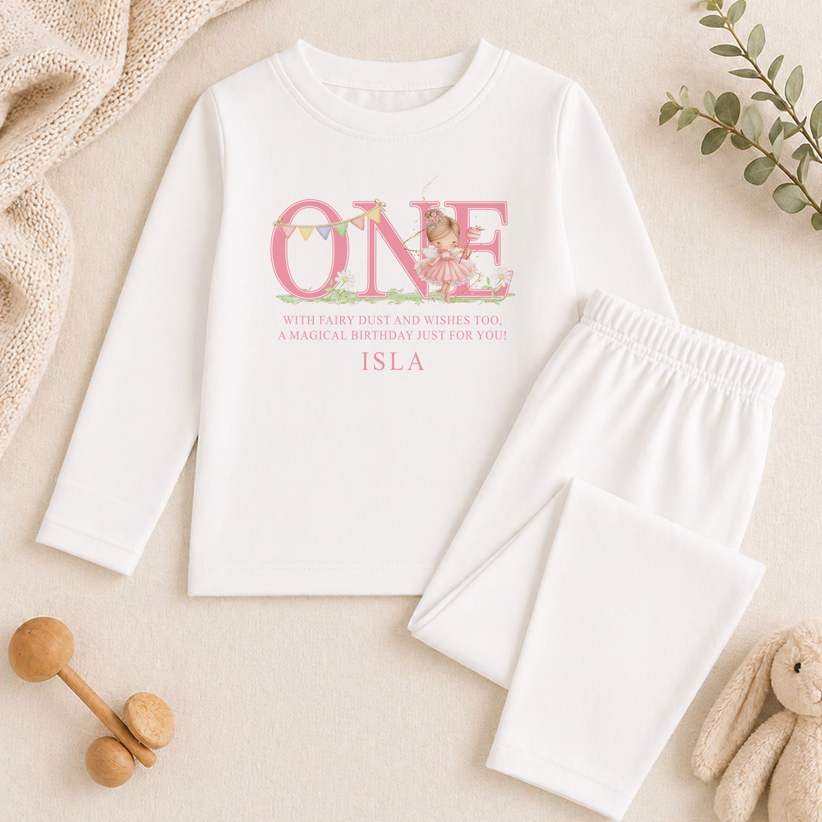 A white long-sleeved shirt with a pink design and text, along with a pair of white pants, a stuffed animal, and some decorative elements, all arranged on a beige background.