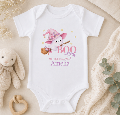 A white baby onesie with a cartoon image of a witch and the text "BOO - TIFAL - My first Halloween - Amelia" printed on it, along with a stuffed bunny and a wooden bib.