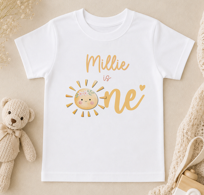 A white t-shirt with a sun graphic and the text "Milly is one" printed on it, along with a teddy bear and other baby items in the background.