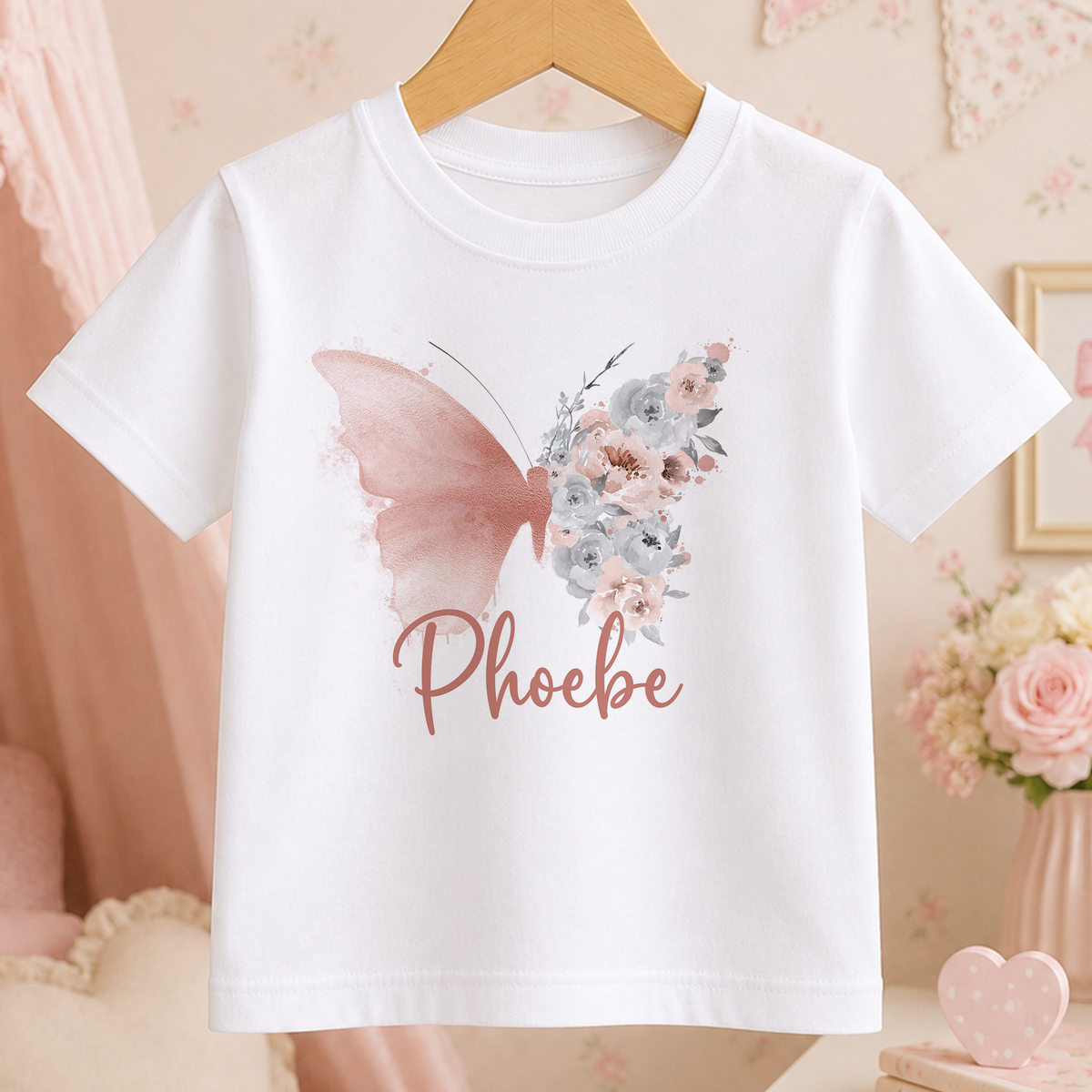 A white t-shirt with a pink butterfly design and the name "Phoebe" printed on it, hanging on a wooden hanger.