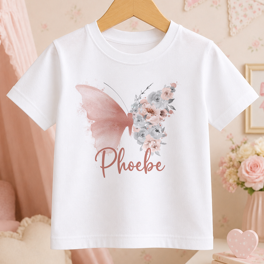 A white t-shirt with a pink butterfly design and the name "Phoebe" printed on it, hanging on a wooden hanger.