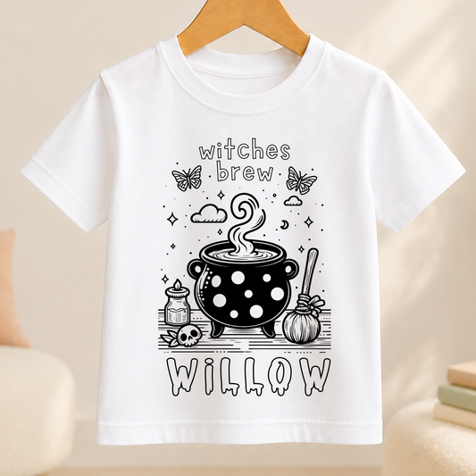 A white t-shirt with a black and white illustration of a cauldron, a witch's hat, and various Halloween-themed elements such as butterflies and skulls.