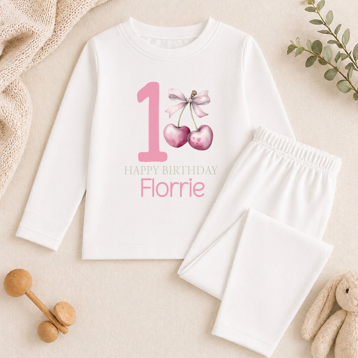 A white onesie with a pink number 1 and a bow, a pair of white pajama bottoms, and a stuffed animal on a beige surface.