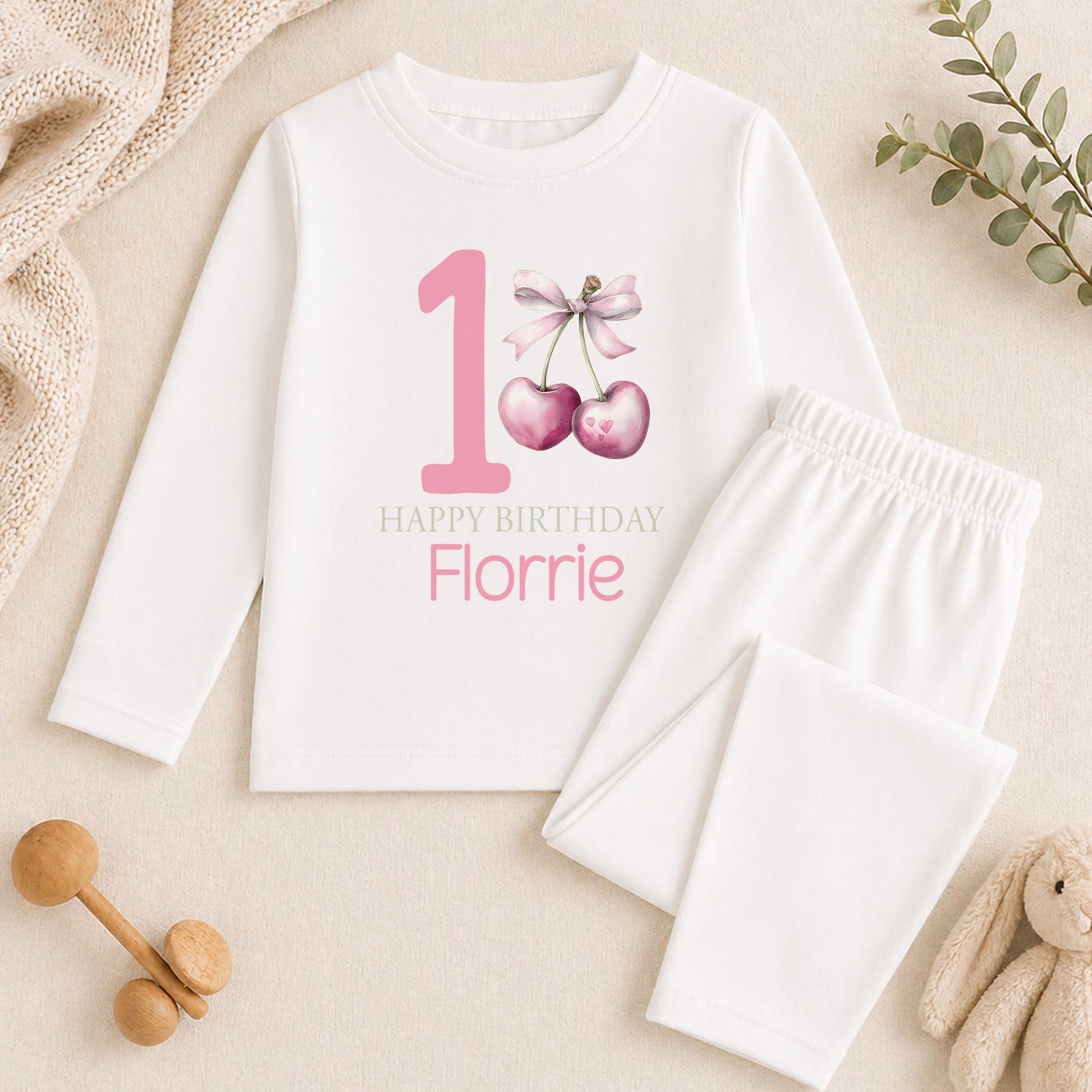 A white onesie with a pink number 1 and a bow, a pair of white pajama bottoms, and a stuffed animal on a beige surface.