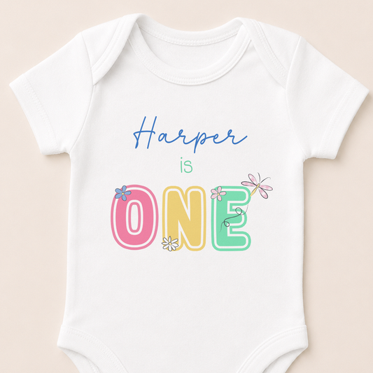 Personalised first birthday vest printed with pastel daisies, a dragonfly and 'is ONE' with your baby's name.