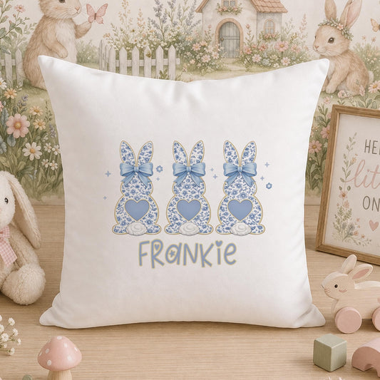 White cushion printed with a design of three blue and white bunny rabbits, hearts and bows, and the personalised name 'Frankie', styled on a wooden surface with nursery toys and a framed picture in the background.