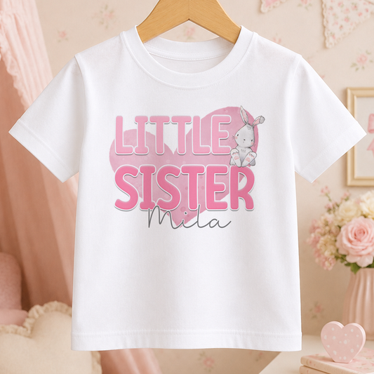 A white t-shirt with the text "LITTLE SISTER Mila" printed on it, along with a small cartoon rabbit.