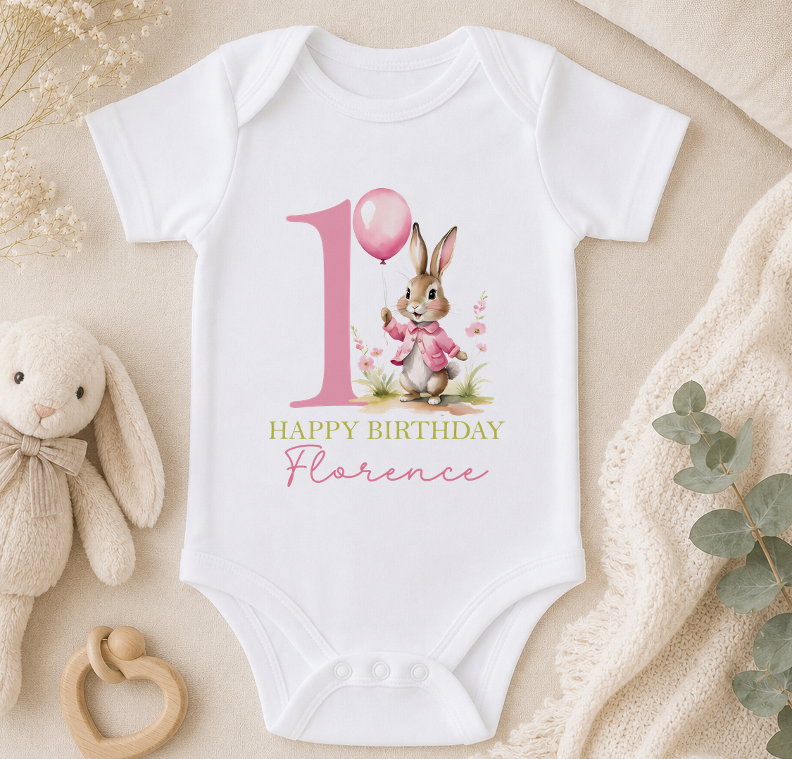 A white onesie with a pink bunny and the number 1, along with the text "Happy Birthday Florence" printed on it, is displayed on a bed surrounded by a stuffed bunny, a teething ring, and some greenery.