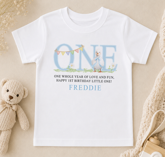 A white t-shirt with a personalized birthday message and a cartoon duck graphic, along with a teddy bear and other baby items.