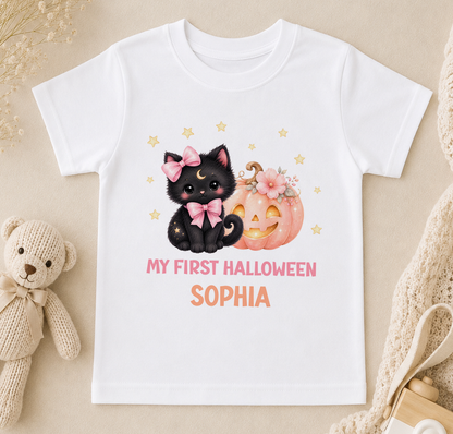 A white t-shirt with a Halloween-themed graphic design featuring a black cat with a bow, a pumpkin, and stars. The text on the t-shirt says "My first Halloween Sophia".