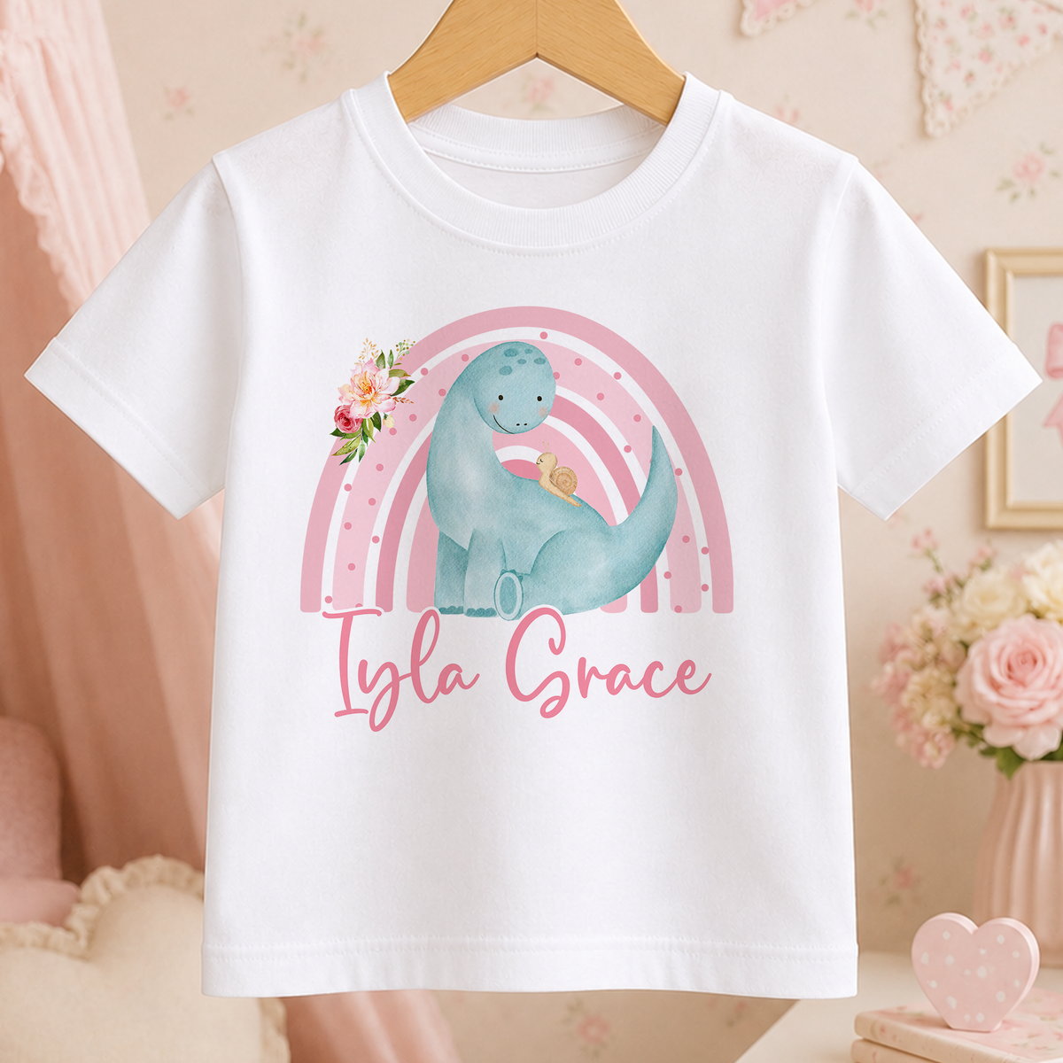 A white t-shirt with a dinosaur graphic and a pink rainbow design, along with the name "Ilya Grace" written on it.