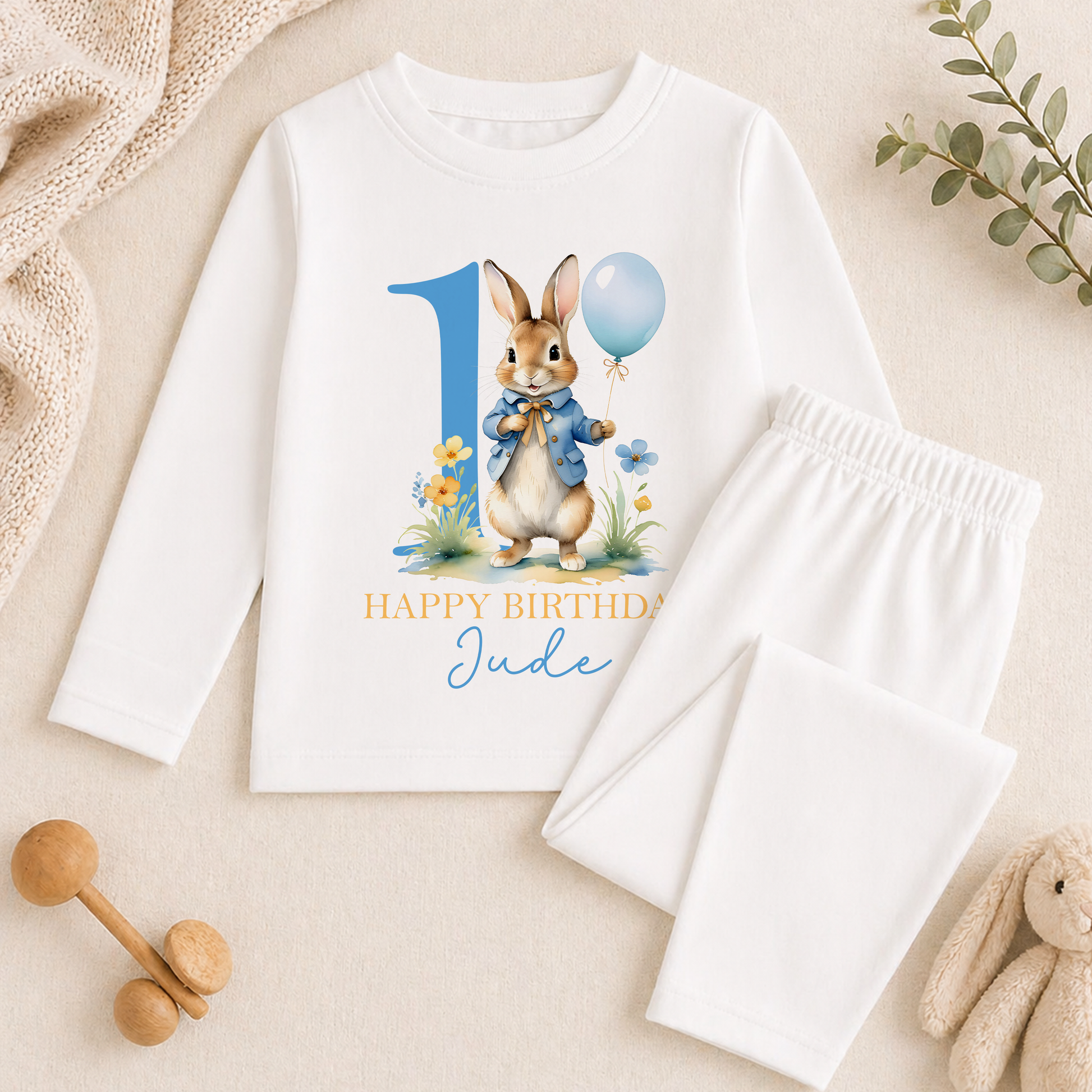 A white long-sleeved shirt with a bunny graphic and the text "Happy Birthday Jude" printed on it, along with a pair of white pajama pants. The shirt and pants are placed on a beige surface, and there are some toys and a plant in the background.