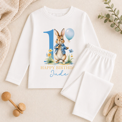 A white long-sleeved shirt with a bunny graphic and the text "Happy Birthday Jude" printed on it, along with a pair of white pajama pants. The shirt and pants are placed on a beige surface, and there are some toys and a plant in the background.