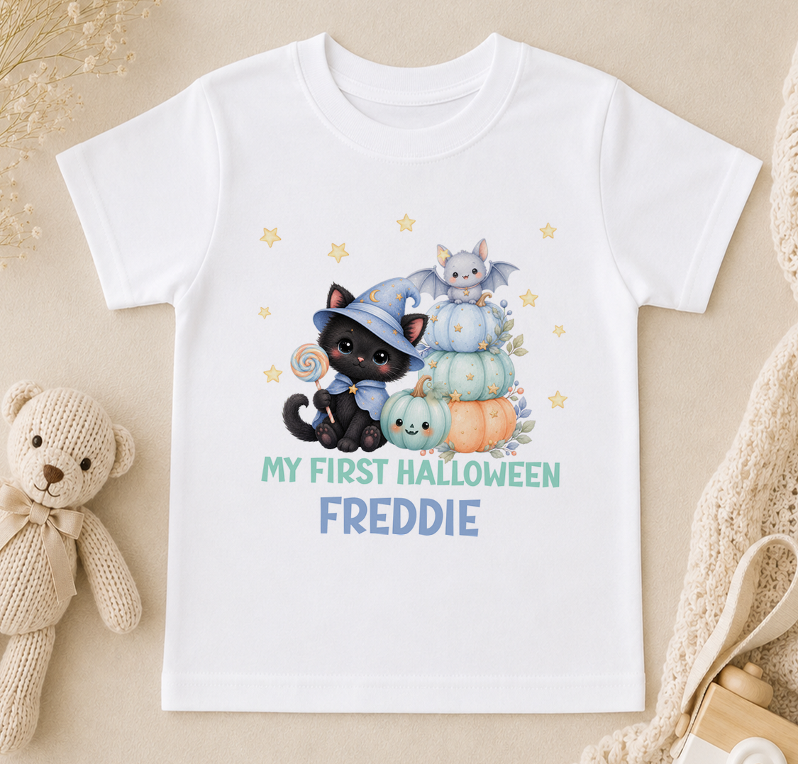 A white t-shirt with a Halloween-themed graphic design featuring a black cat, a witch, and pumpkins, along with the text "My first Halloween Freddie".