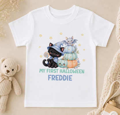 A white t-shirt with a Halloween-themed graphic design featuring a black cat, a witch, and pumpkins, along with the text "My first Halloween Freddie".