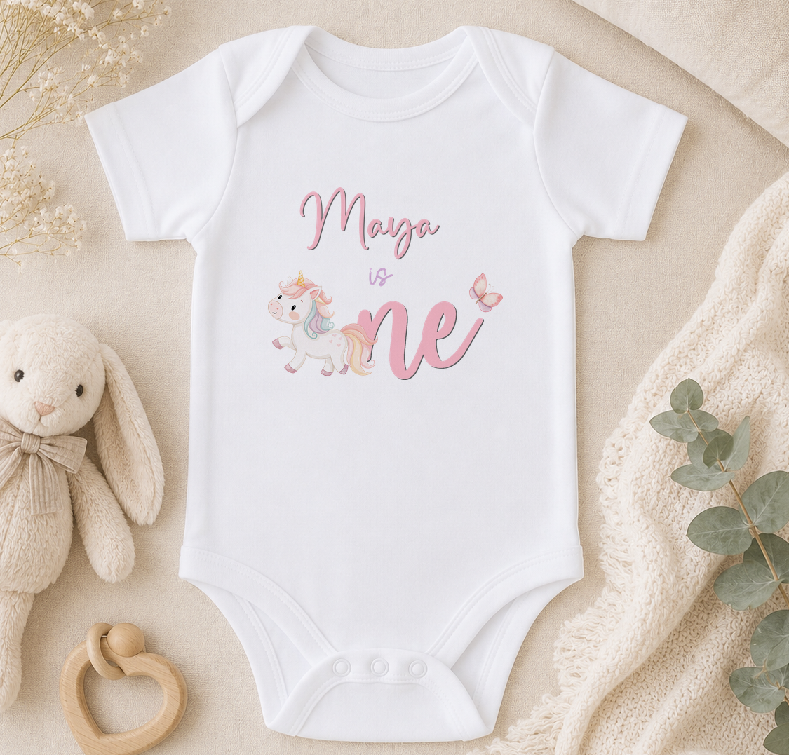 A white onesie with a unicorn and the text "Maya is one" printed on it, along with a stuffed bunny and a wooden bib.