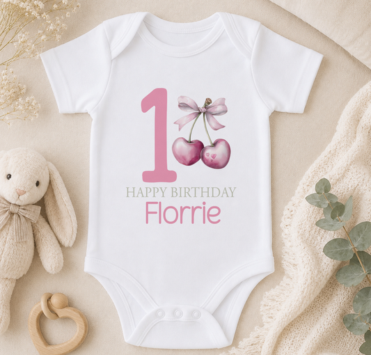 A white onesie with a pink number 1 and a bow, along with a stuffed animal and some greenery, arranged on a beige surface.