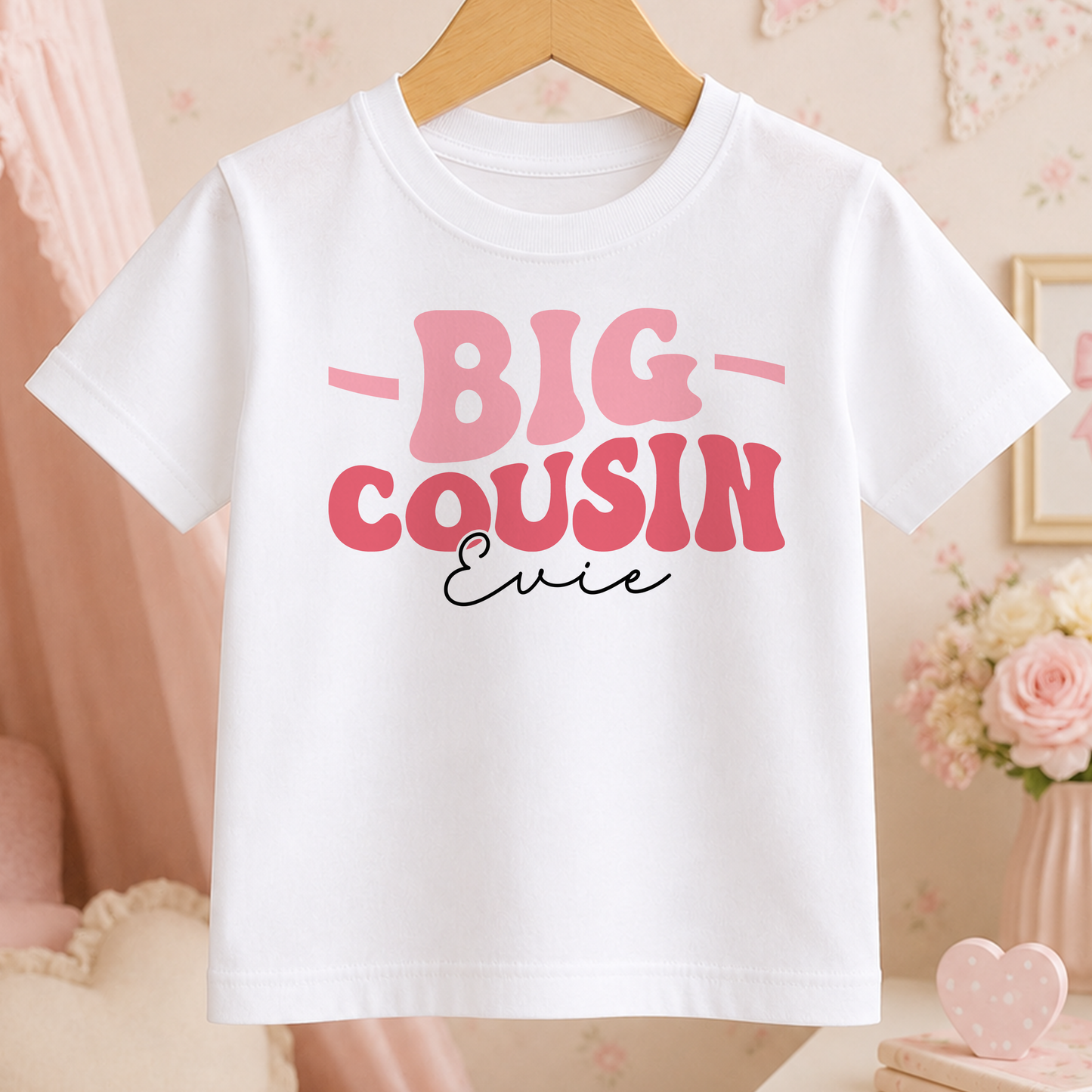 A white t-shirt with pink text that says "BIG COUSIN Ernie" hanging on a wooden hanger.