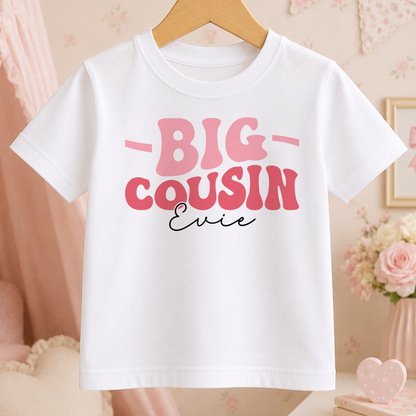 A white t-shirt with pink text that says "BIG COUSIN Ernie" hanging on a wooden hanger.