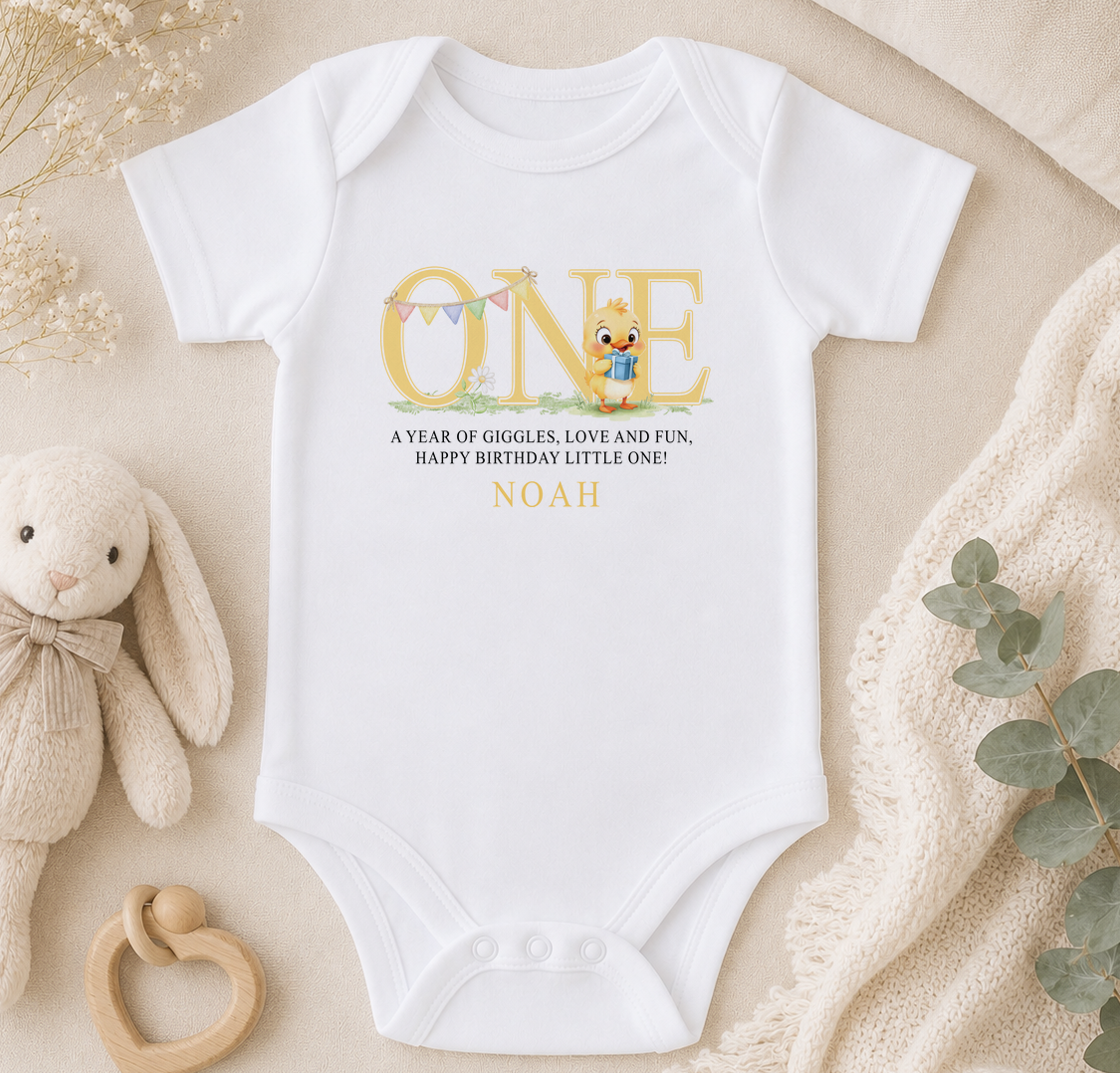 A white onesie with a cartoon character and text, placed on a beige surface with a stuffed animal and greenery.