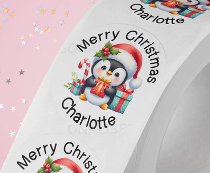Glossy white round Christmas sticker personalised with a name, printed with a cute cartoon penguin design, shown on a strip of stickers