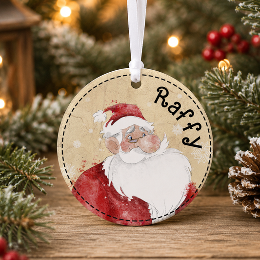 A round ornament featuring a cartoon Santa Claus wearing a red suit and hat, with the name "Raffy" written on it. The ornament is hanging from a string, and there are pine branches and red berries in the background.