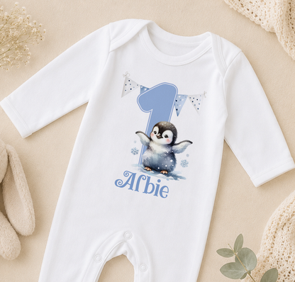 A white onesie with a penguin design and the number 1, along with the text "Alfie" printed on it, is displayed on a beige surface.