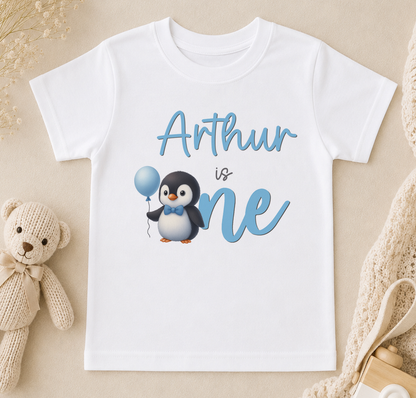 A white t-shirt with a penguin and a balloon, along with the text "Arthur is one" printed on it.