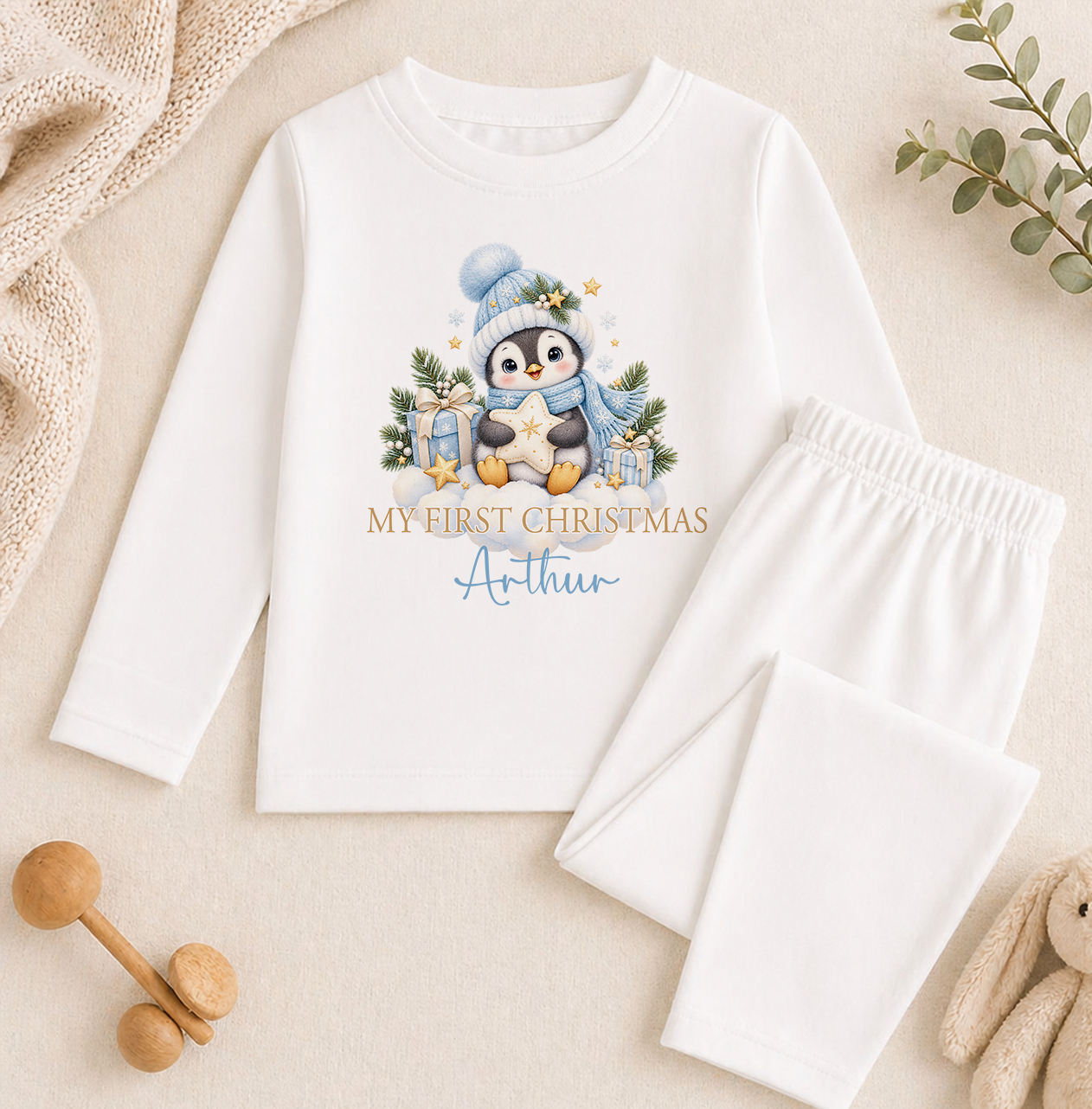 A white long-sleeved shirt with a penguin graphic and the text "My first Christmas Arthur" printed on it, along with a pair of white pajama pants. The shirt and pants are placed on a beige surface, and there are some wooden toys and greenery in the background.