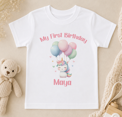 A white t-shirt with a printed design of a unicorn and balloons, along with the text "My First Birthday Maya".