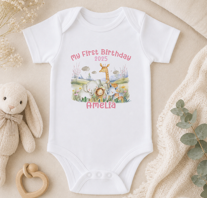 A white onesie with a personalized birthday message and an illustration of animals, including a giraffe, elephants, and a rabbit, is displayed on a bed surrounded by stuffed animals and a blanket.