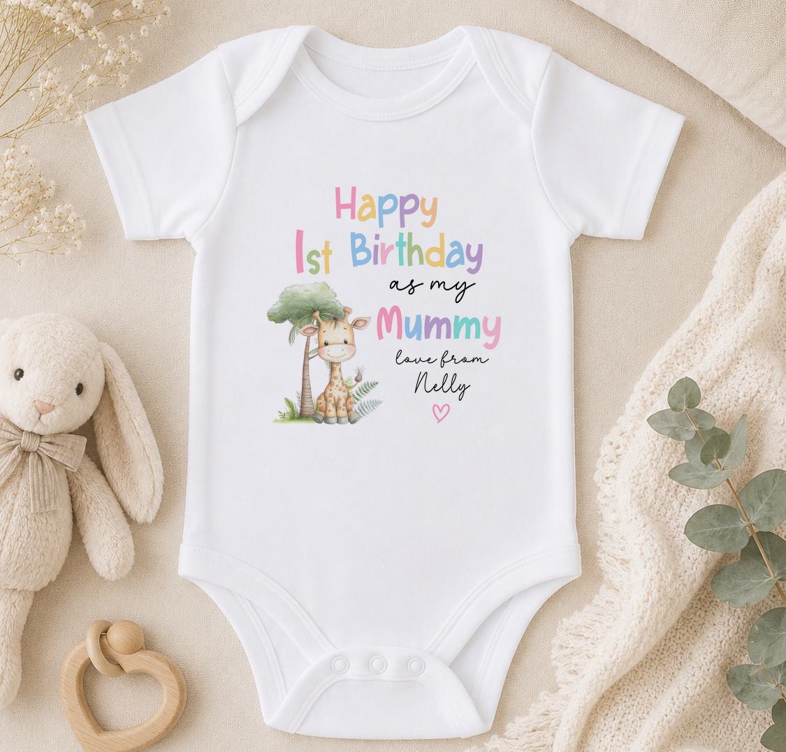 A white onesie with a giraffe and text that reads "Happy 1st Birthday as my Mummy love from Nelly" is displayed on a beige surface, accompanied by a stuffed animal and greenery.