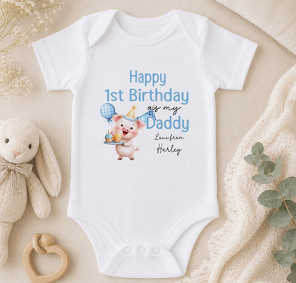 A white onesie with a cartoon pig and text that reads "Happy 1st Birthday as my Daddy Harley" is displayed on a beige surface, accompanied by a stuffed animal and some greenery.