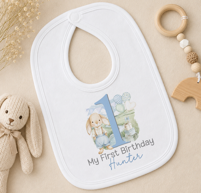 A white baby bib with a blue number 1 and the text "My First Birthday Hunter" printed on it, along with a small stuffed rabbit and a wooden bead ringer.