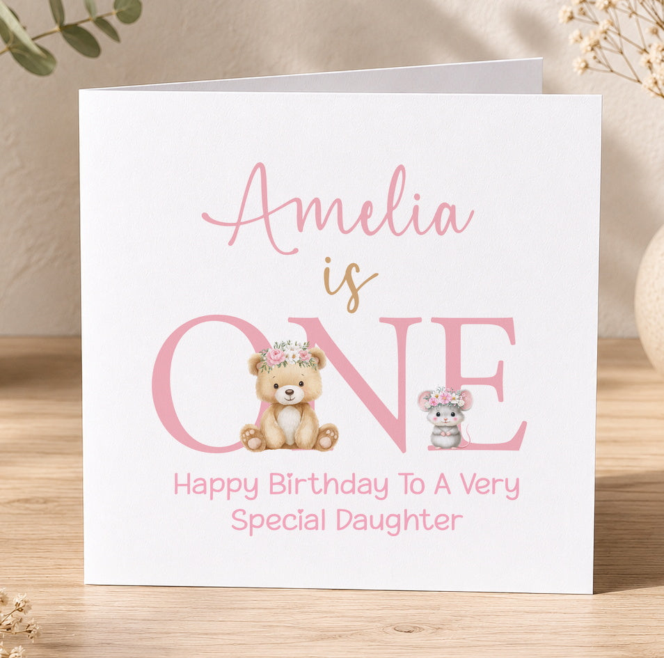 Pink 1st birthday card for a girl featuring a floral crown, teddy bear and mouse design with the message 'Amelia is ONE – Happy Birthday to a Very Special Daughter'.