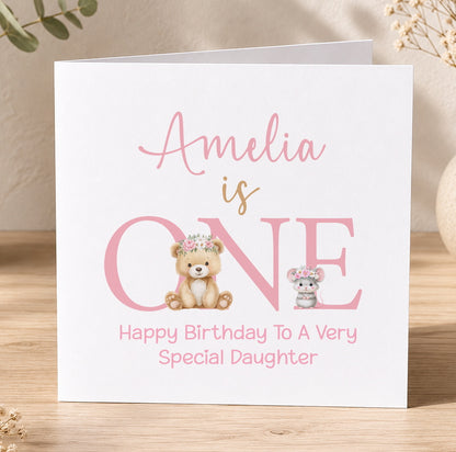 Pink 1st birthday card for a girl featuring a floral crown, teddy bear and mouse design with the message 'Amelia is ONE – Happy Birthday to a Very Special Daughter'.