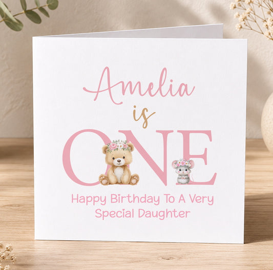 Pink 1st birthday card for a girl featuring a floral crown, teddy bear and mouse design with the message 'Amelia is ONE – Happy Birthday to a Very Special Daughter'.