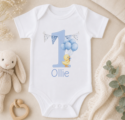 A white onesie with a blue duck and balloons, along with a stuffed animal and a blanket, arranged on a surface.