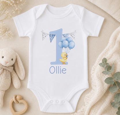 A white onesie with a blue duck and balloons, along with a stuffed animal and a blanket, arranged on a surface.