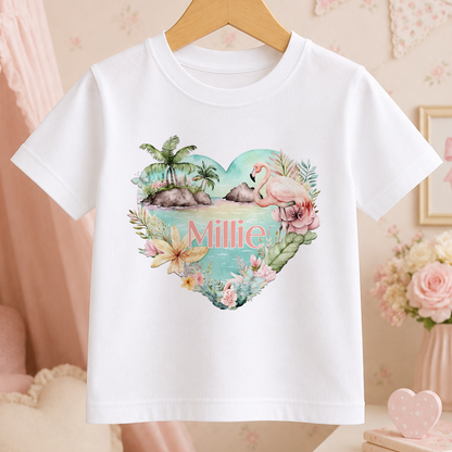 A white t-shirt with a heart-shaped design featuring a flamingo, palm trees, and flowers, along with the name "Millie" written on it.