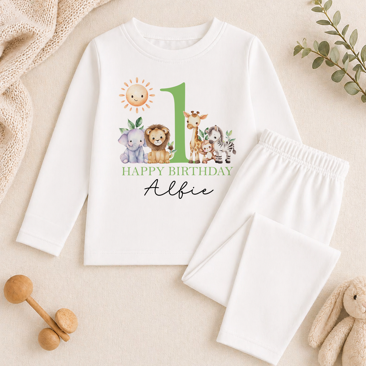 Long-sleeved top and white trousers from the safari friends birthday set, printed with jungle animals and 'Happy Birthday Albie', styled with baby toys and greenery.