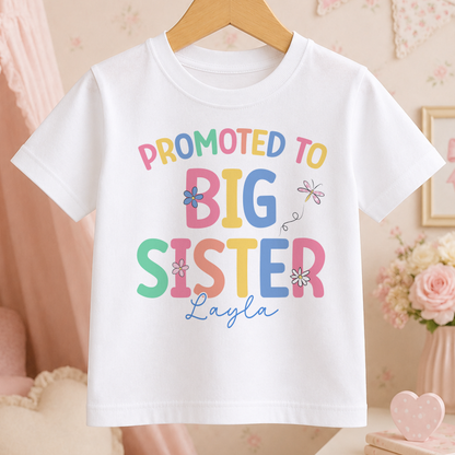 A white t-shirt with a colorful text that reads "PROMOTED TO BIG SISTER Layla" printed on it.
