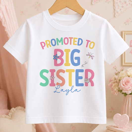 A white t-shirt with a colorful text that reads "PROMOTED TO BIG SISTER Layla" printed on it.