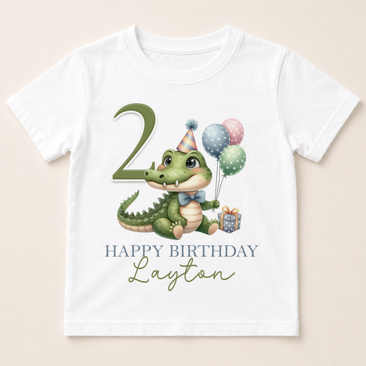 A white t-shirt with a cartoon image of a green alligator wearing a party hat and holding balloons, along with the text "Happy Birthday Layton" printed on it.