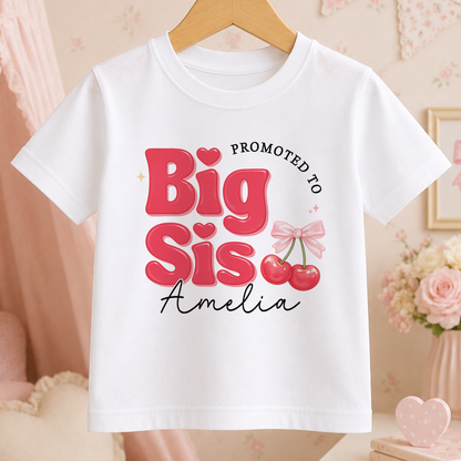 A white t-shirt with a pink and red design that says "Promoted to Big Sister Amelia" and features a bow and cherries.